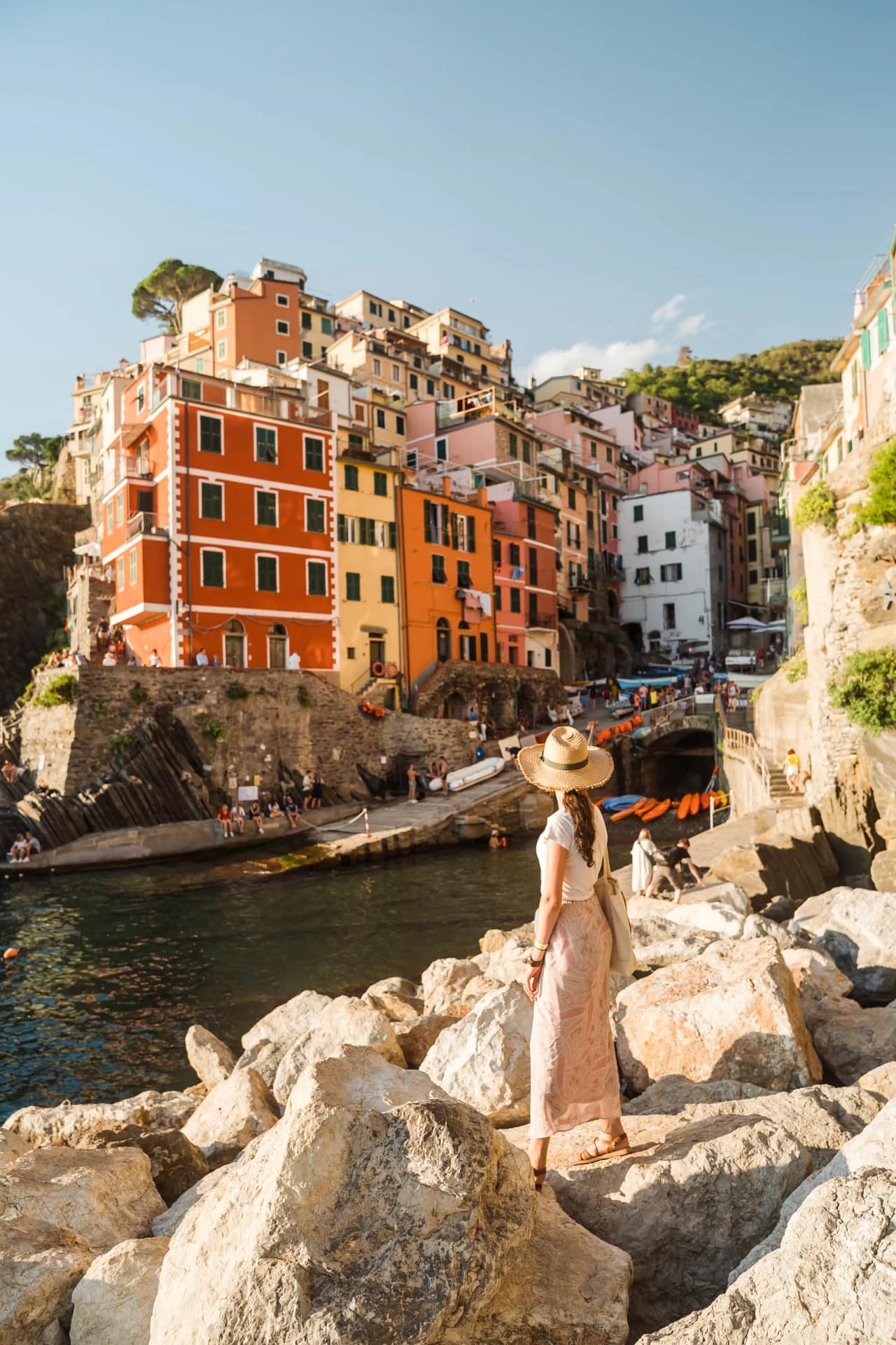 Golden hour view over the colorful harbor of Riomaggiore, Cinque Terre, Italy Woman wearing a straw hat and long skirt standing on the rocky breakwater, admiring the vibrant red and yellow houses of Riomaggiore village in the Cinque Terre, Italian Riviera, during sunset.